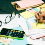 Person writing notes on financial documents with calculator and paperwork, budgeting and tax planning workspace.