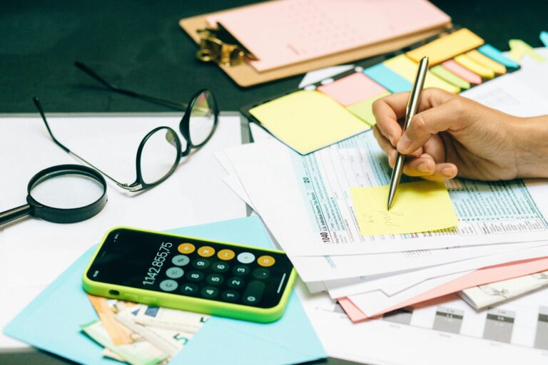Person writing notes on financial documents with calculator and paperwork, budgeting and tax planning workspace.